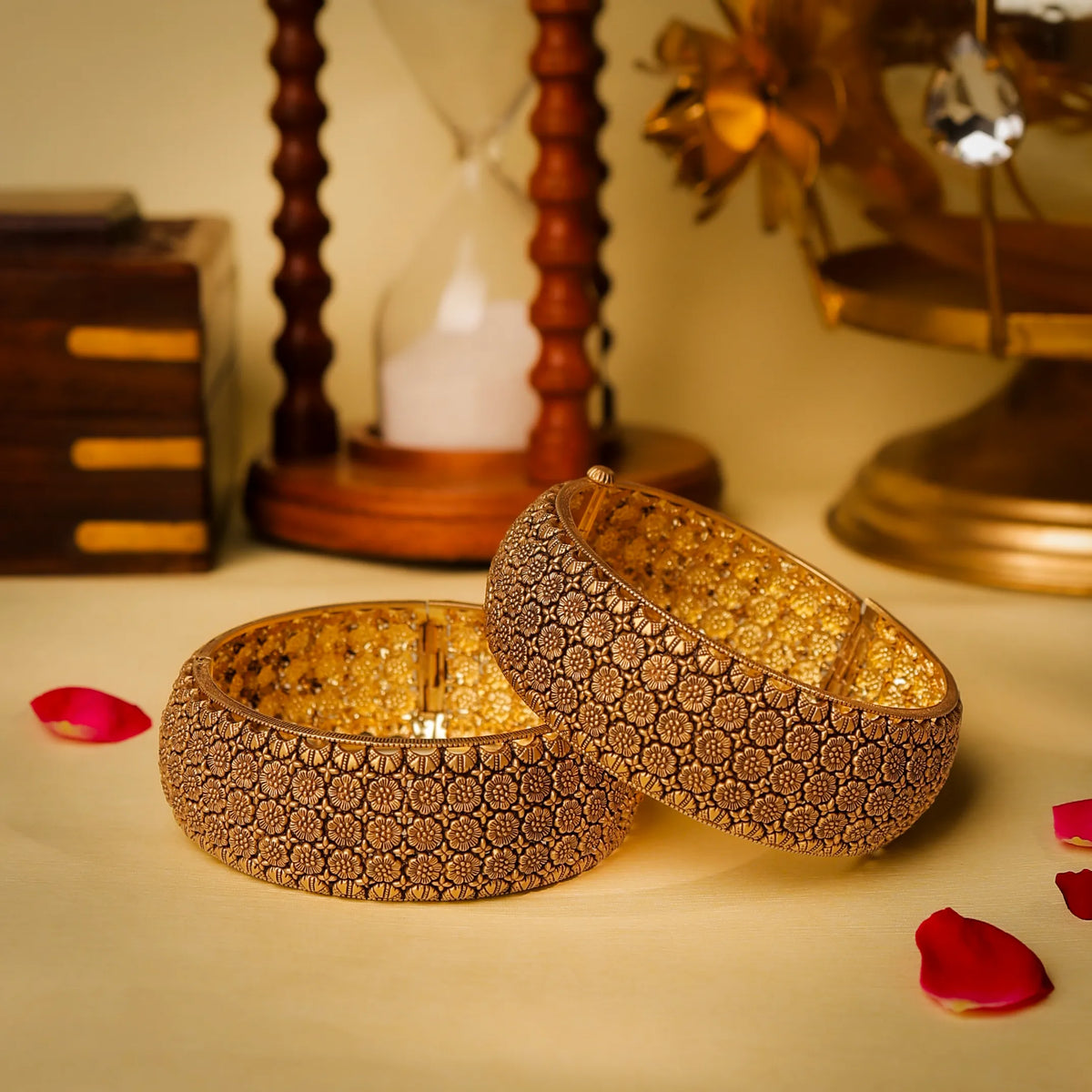 Two gold bangles with intricate patterns on a decorative surface with rose petals.