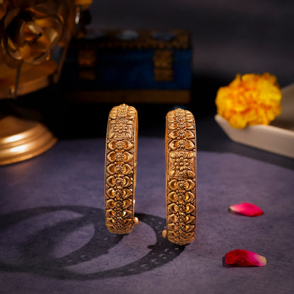 Pair of gold bangles with intricate designs on a dark surface with flowers.